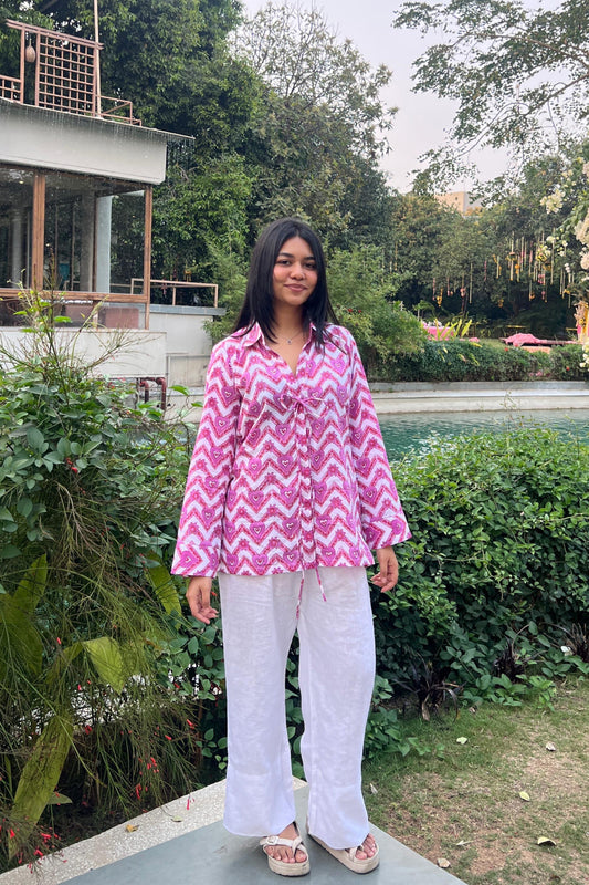 Person wearing a pink and white zigzag patterned shirt with white pants standing in a garden.