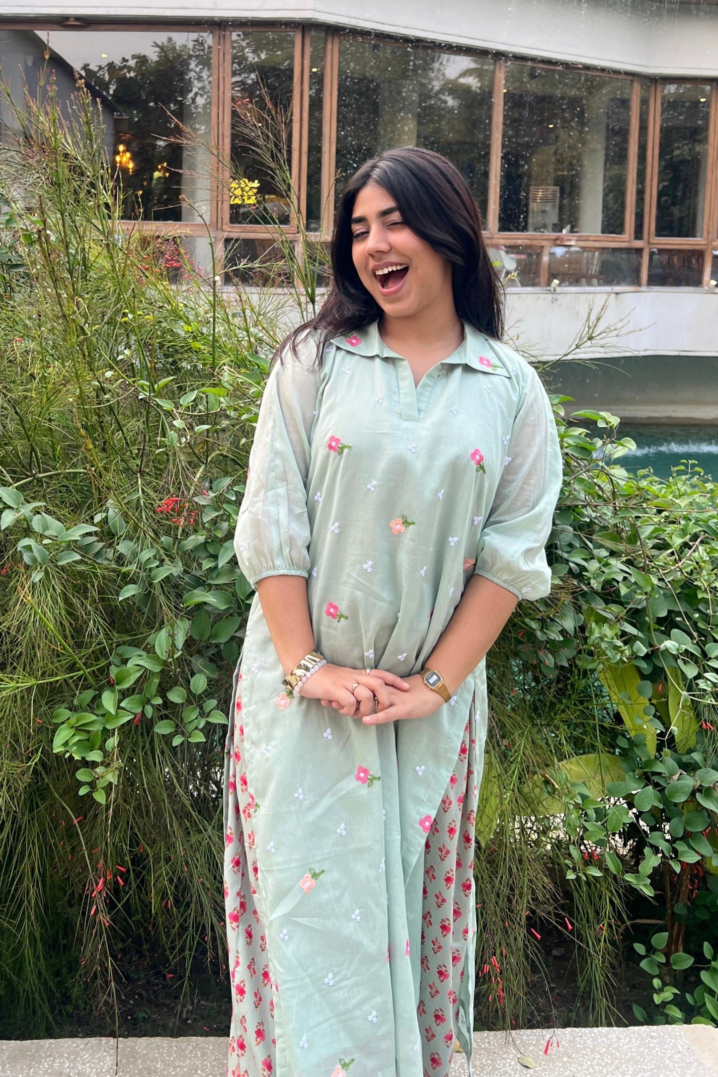 Woman in a light green dress with floral patterns standing outdoors with greenery in the background