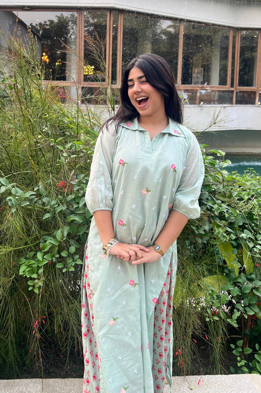 Woman in a light green dress with floral patterns standing outdoors with greenery in the background