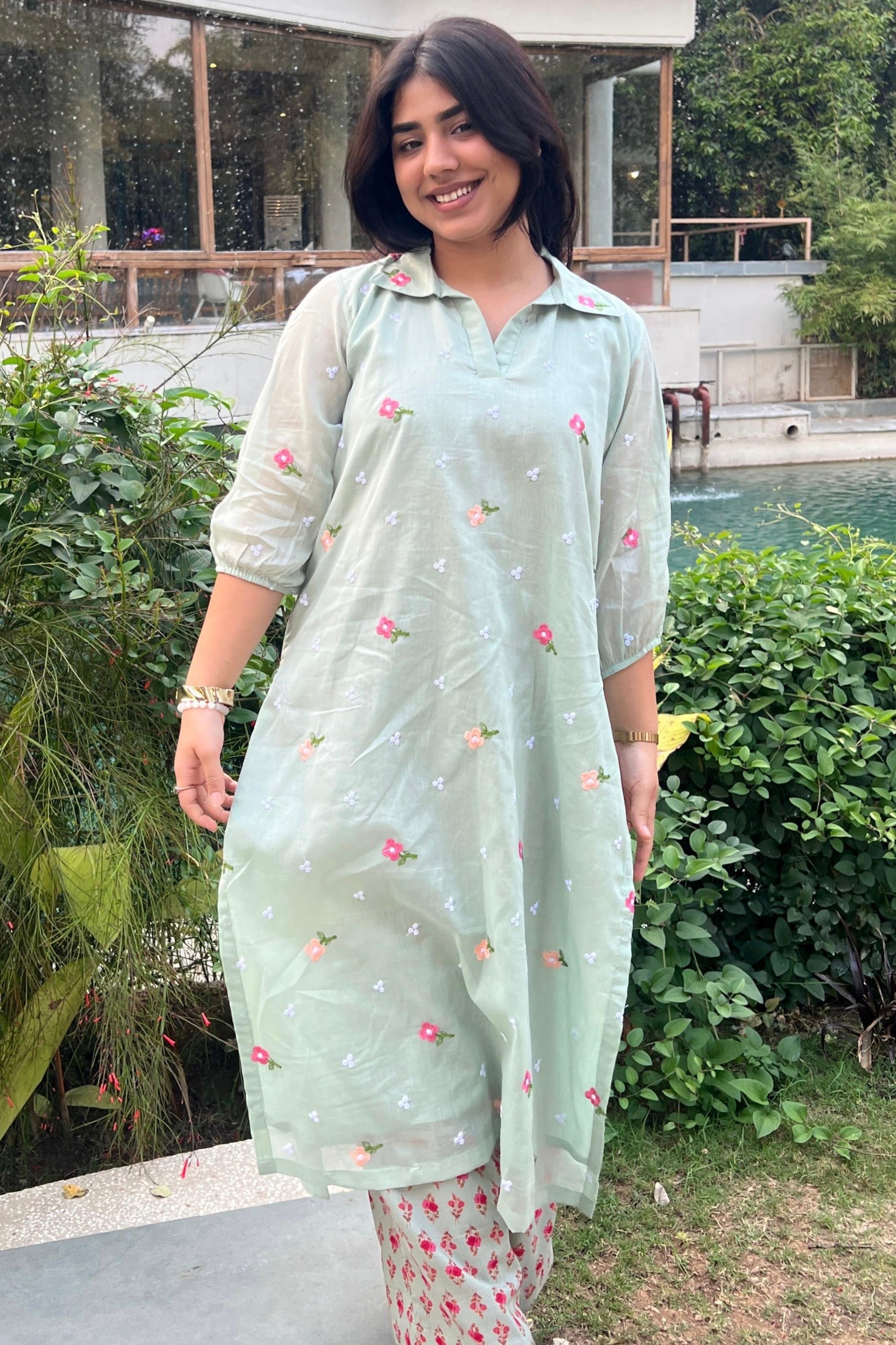 Woman in a light green traditional outfit with floral patterns standing outdoors by a pool.